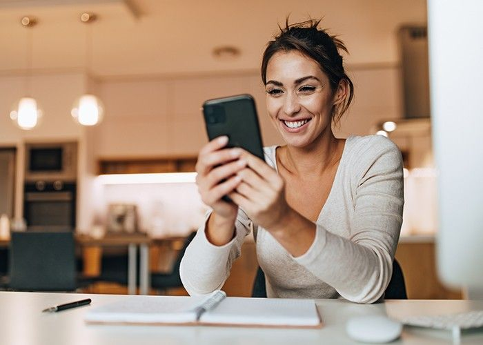 Woman sitting at her desk, smiling while holding her phone with two hands.